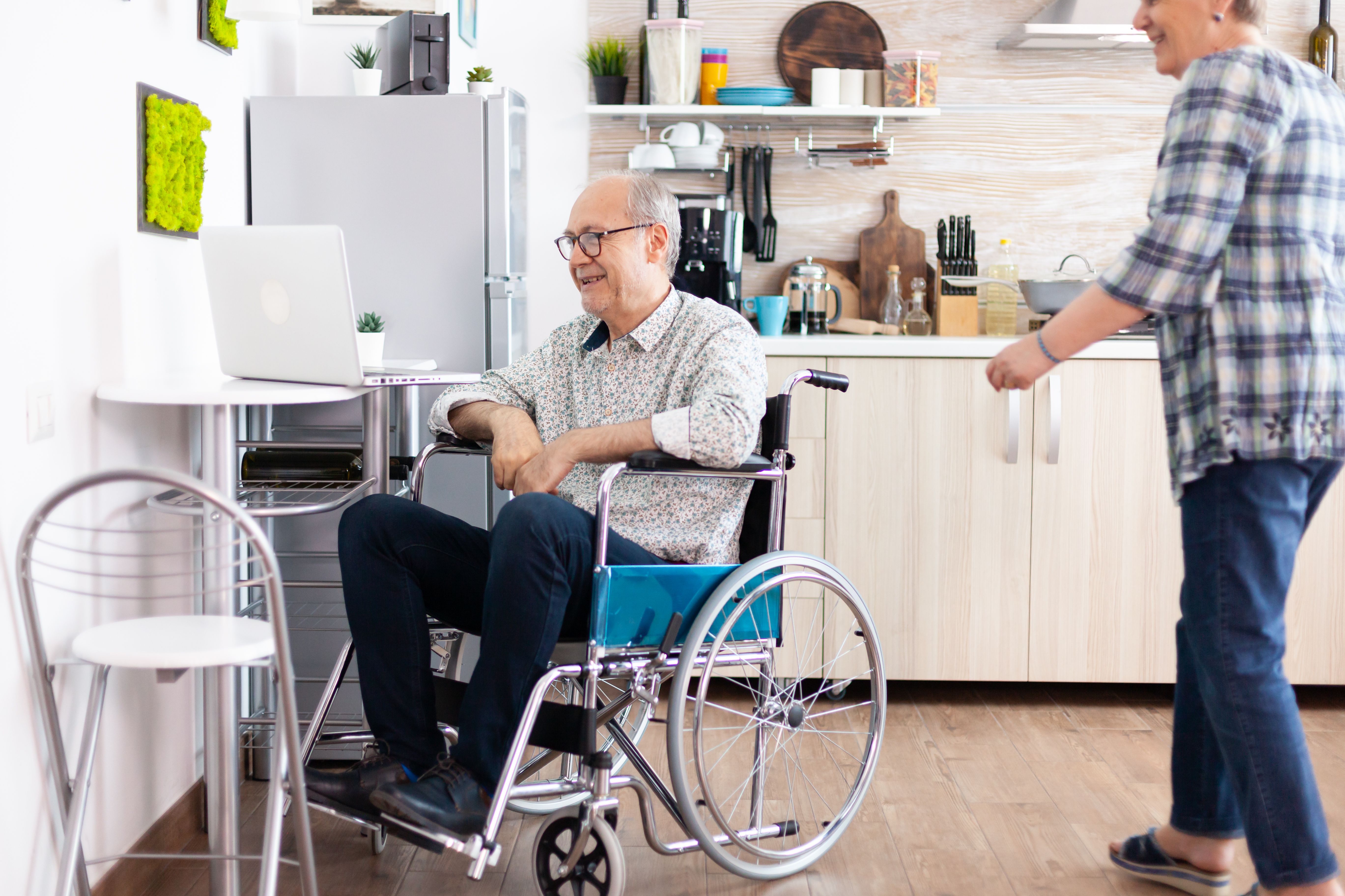 senior couple laughing using laptop husband calling his wife near him video call with grandchildrens sitting kitchen paralyzedhandicapped old elderly man using communication techonolgy
